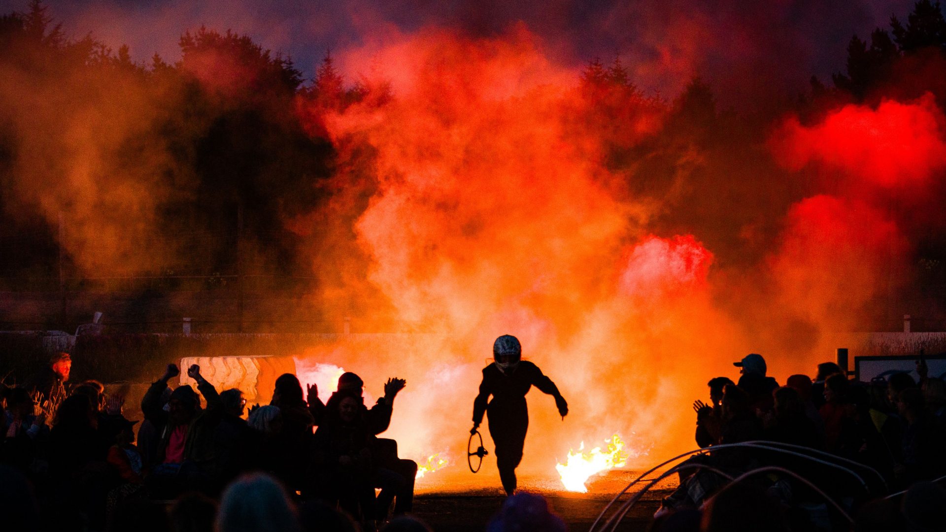 A raceway driver runs out of the smoke and fire wearing overalls and a racing helmet. Kneebone Cadillac by Wildworks
