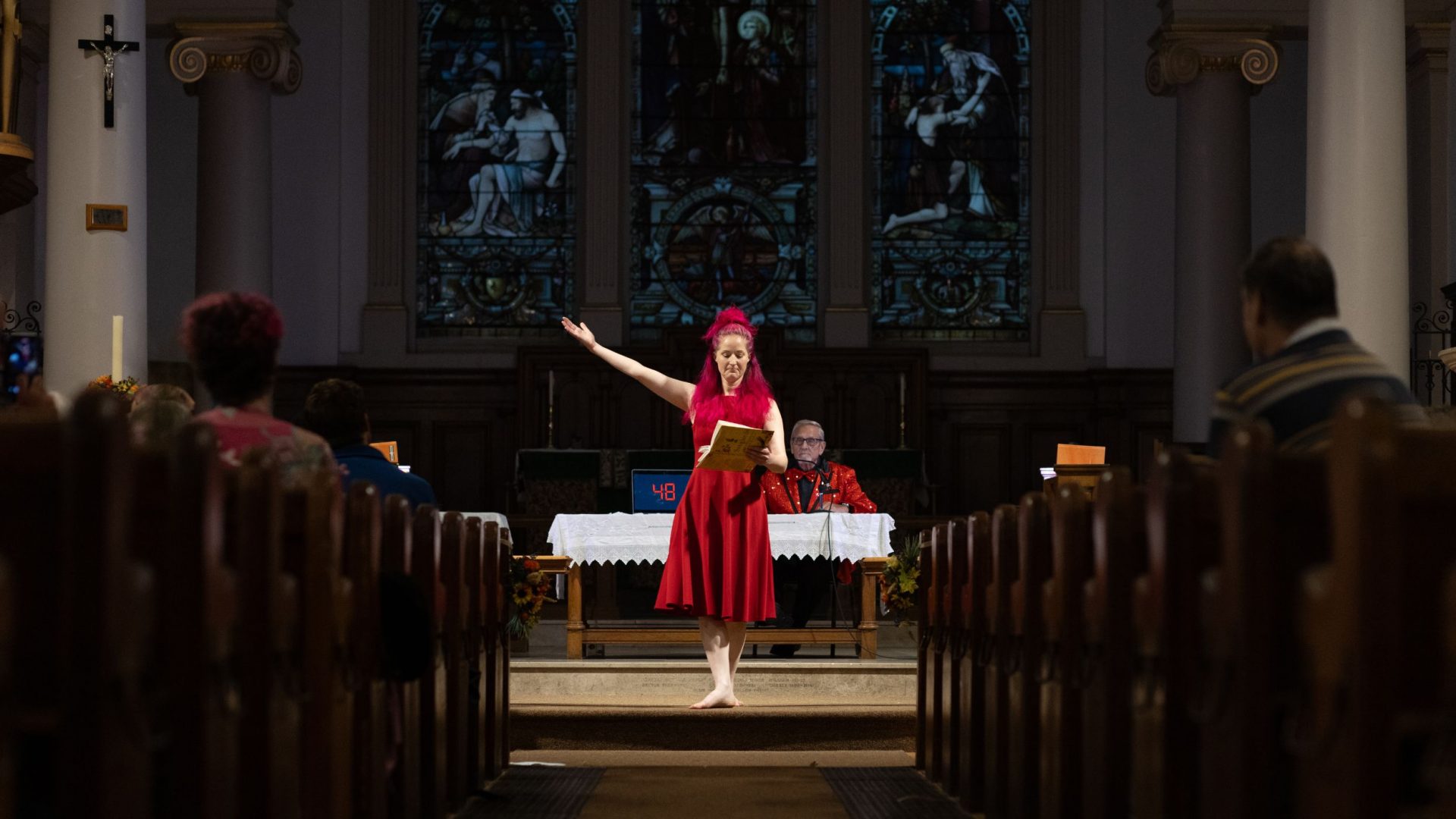 A performer in a red dress stands at the front of a church in front of stained glass windows.