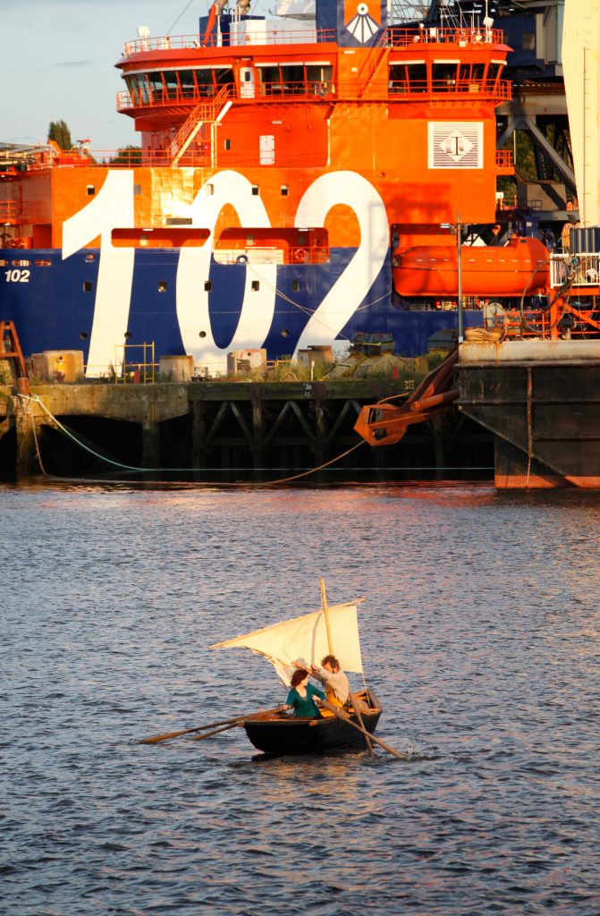 A tiny ship sails on the River Tyne. In the background is a very large ship. A Beautiful Journey by Wildworks