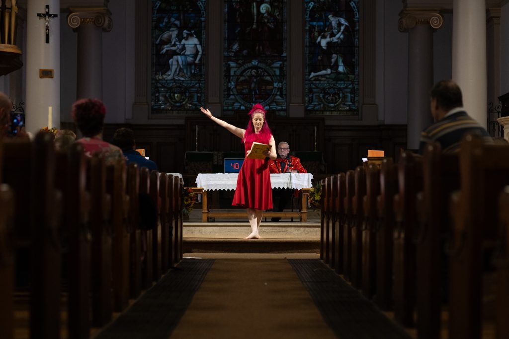 Performer satands in a red dress and hat at the front of a church