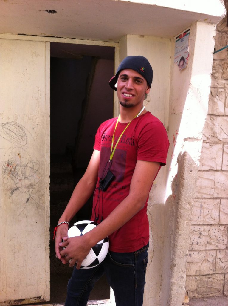 Mohammed is wearing a red shirt and a backwards baseball cap and holding a football. Nablus, City of Stories by Wildworks