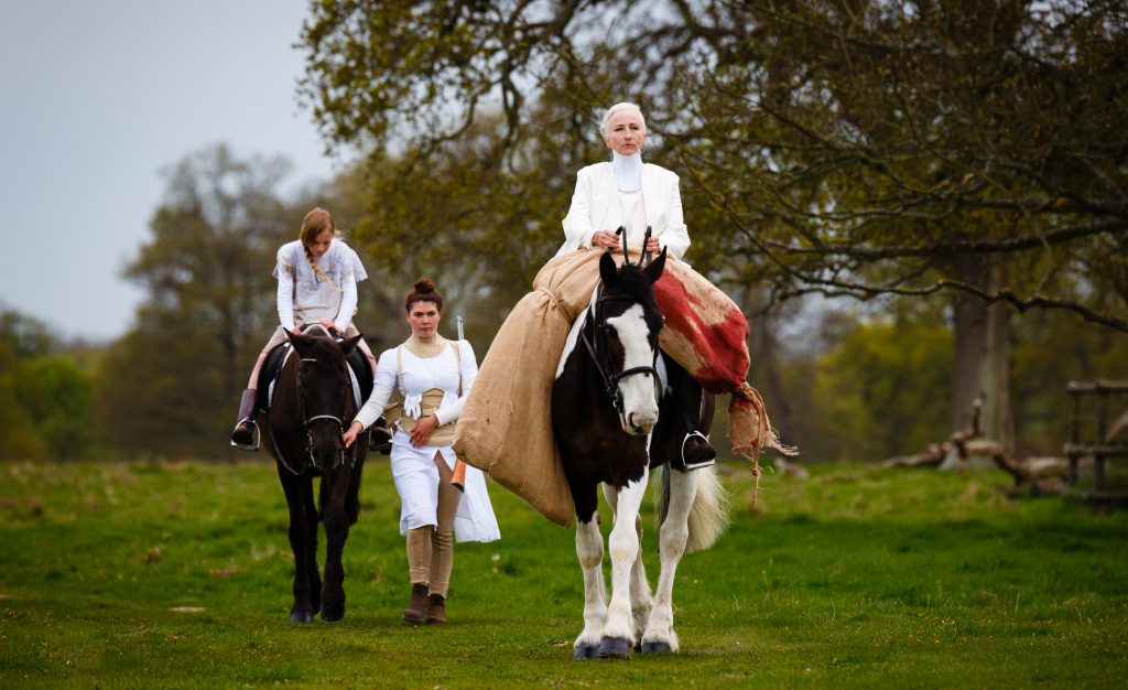 Mother riding a magnificent shire horse and carrying the body of a bleeding wolf across the saddle, surrounded by fiercely corseted maids. 