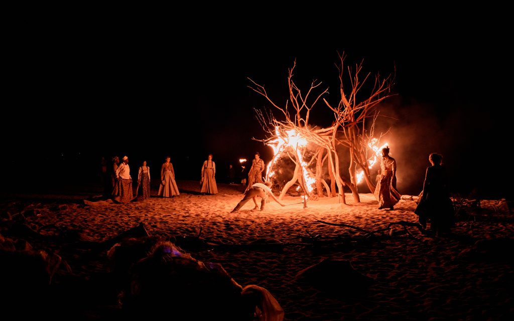 Performers circle the tree at the water's edge. It is on fire. I am Kevin by Wildworks