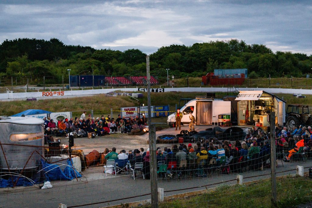 The set of The Kneebone Cadillac Set in the middle of United Downs Raceway. An audience is watching. 