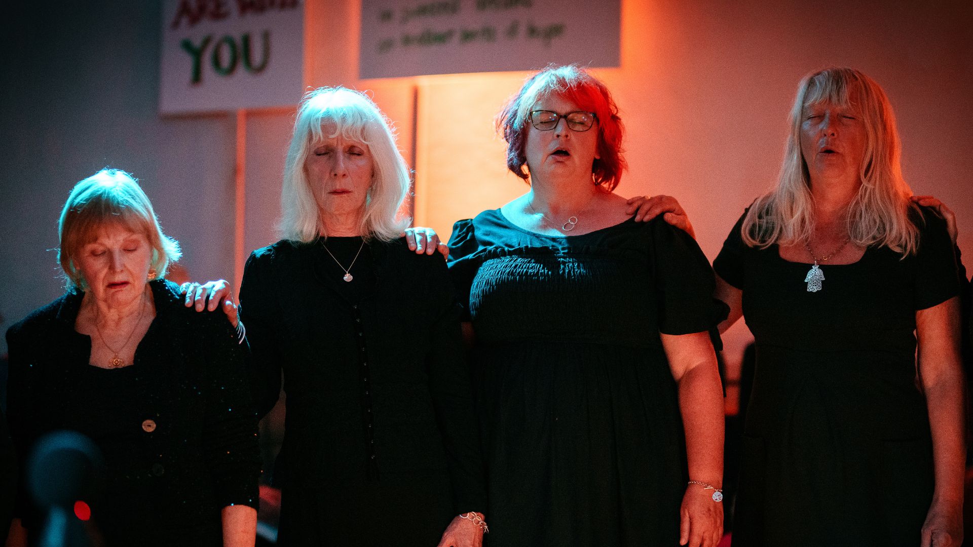 Four women stood in a line. Eye closed, singing during From West Cornwall to the West Bank by Wildworks.