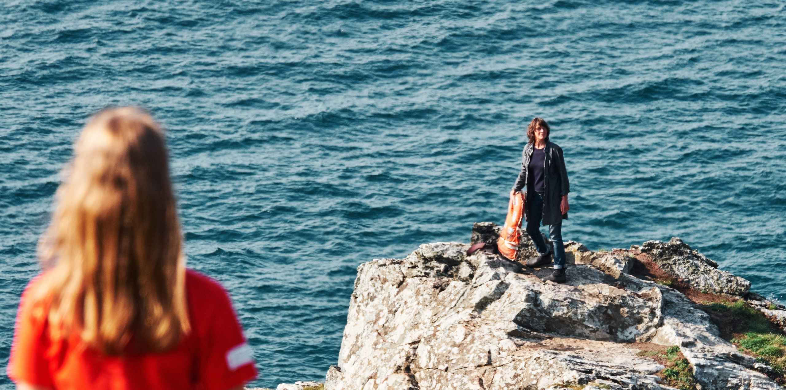 A women stands on the endge of a cliff with the ocean behind her. A girl looks out at her.