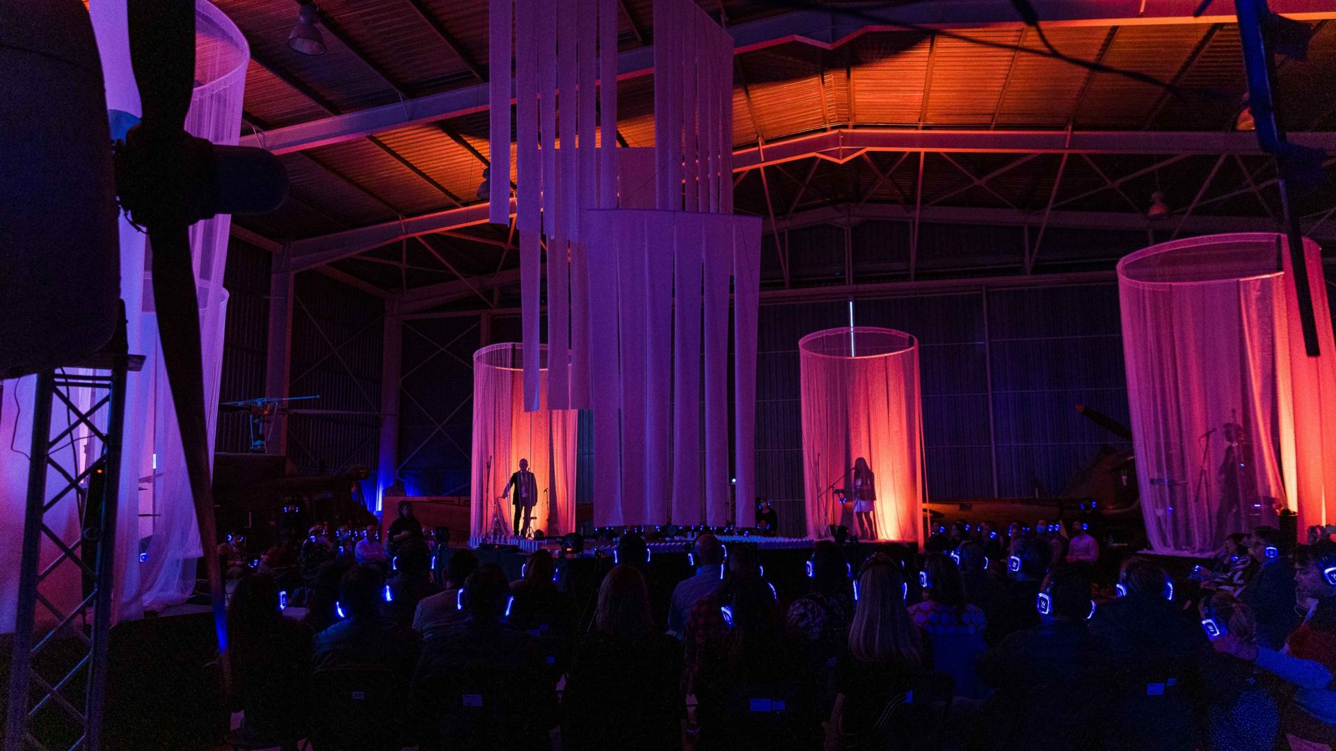 Arrow of time set in Malta Aviation Museum in Ta' Qali with Wildworks and Etnika, Magna Żmien, Buena Sombra Films Two stages are lit up with performers.