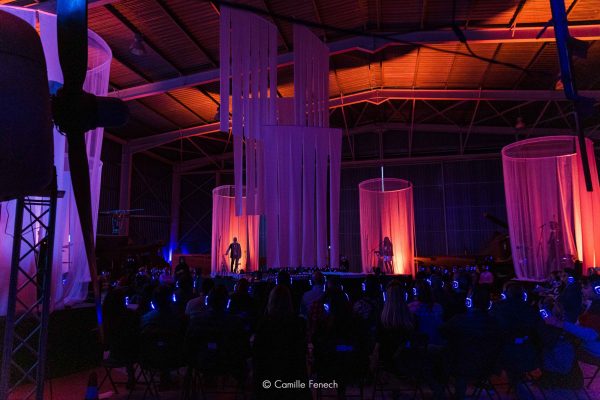 Two performers lit on the stage inside Malta Aviation Museum in Ta' Qali. The audience are wearing headphones. Arrow of Time by Wildworks