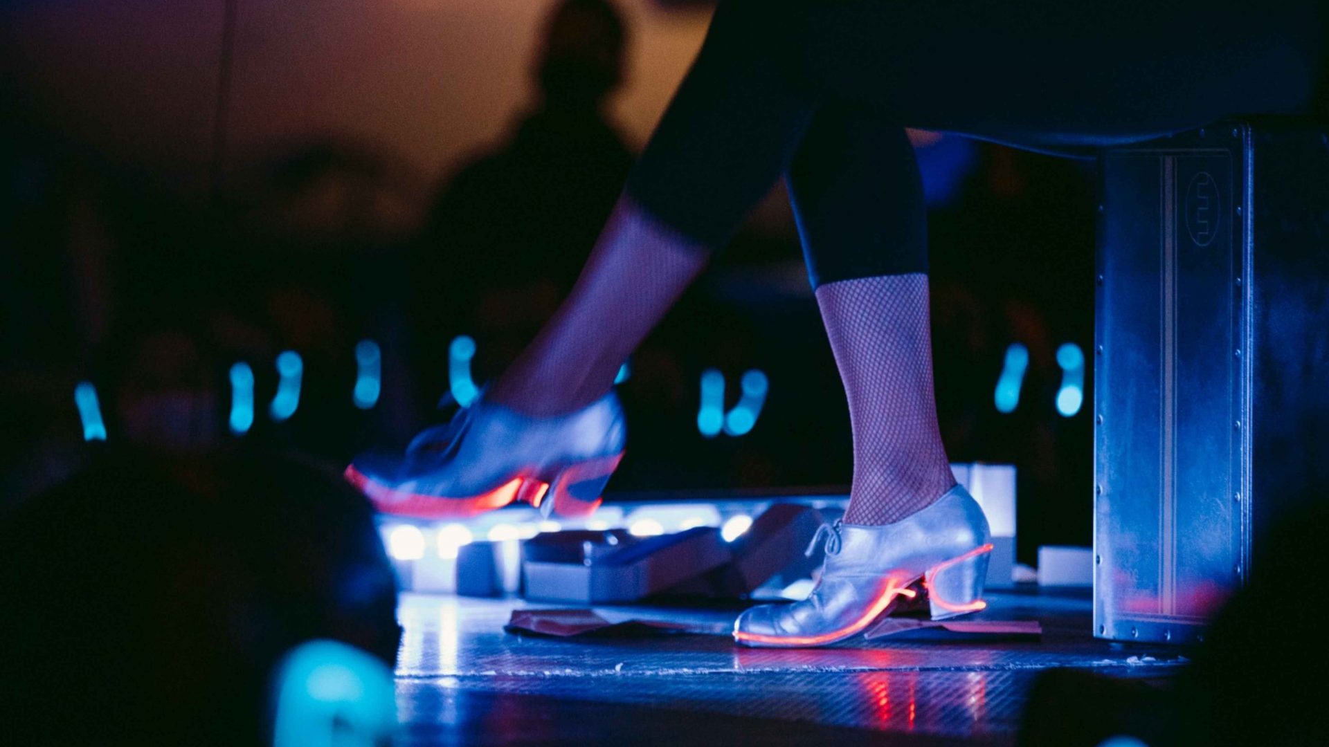 Shot of someone's legs. They are wearing silver heeled shoes and sat on a stage. Arrow of time set in Malta Aviation Museum in Ta' Qali with Wildworks and Etnika, Magna Żmien, Buena Sombra Films