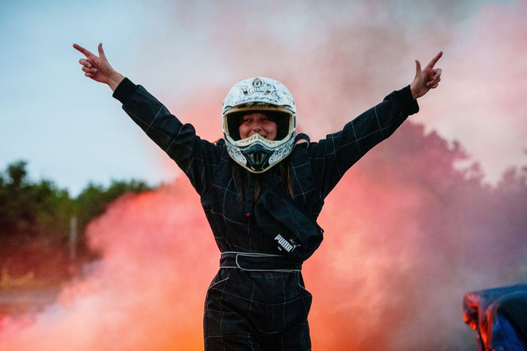 Woman with hands in the air coming out of smoke, racing helmet on, she is smiling. Kneebone Cadillac by Wildworks