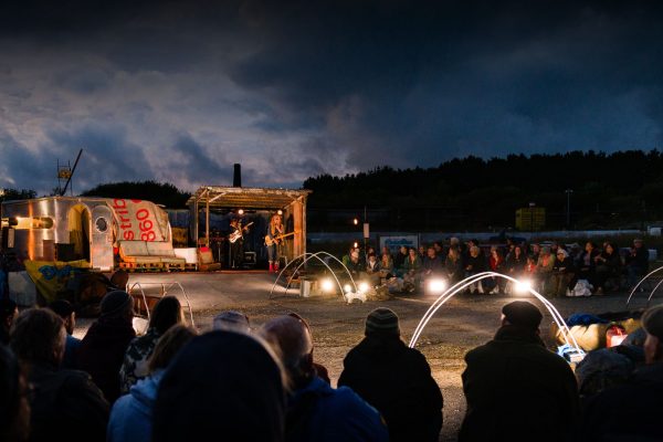 The Kneebone Cadillac set. A carvan stands next to a stage with musicians playing guitars. Set against a backdrop of Cornish tin mine at dusk. The audience is in the foreground.