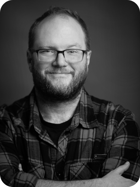 Black and white headshot of Carl Grose. Carl is wearing a checked shirt and wearing glasses.