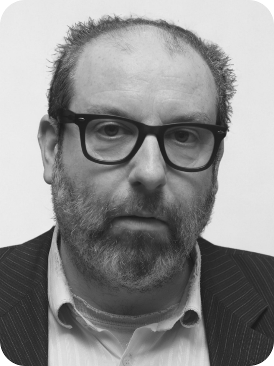 Black and white headshot of Ed Gaughan. He wears thick rimmed black sqaure glasses, he's wearing a shirt and jacket. He has a beard.