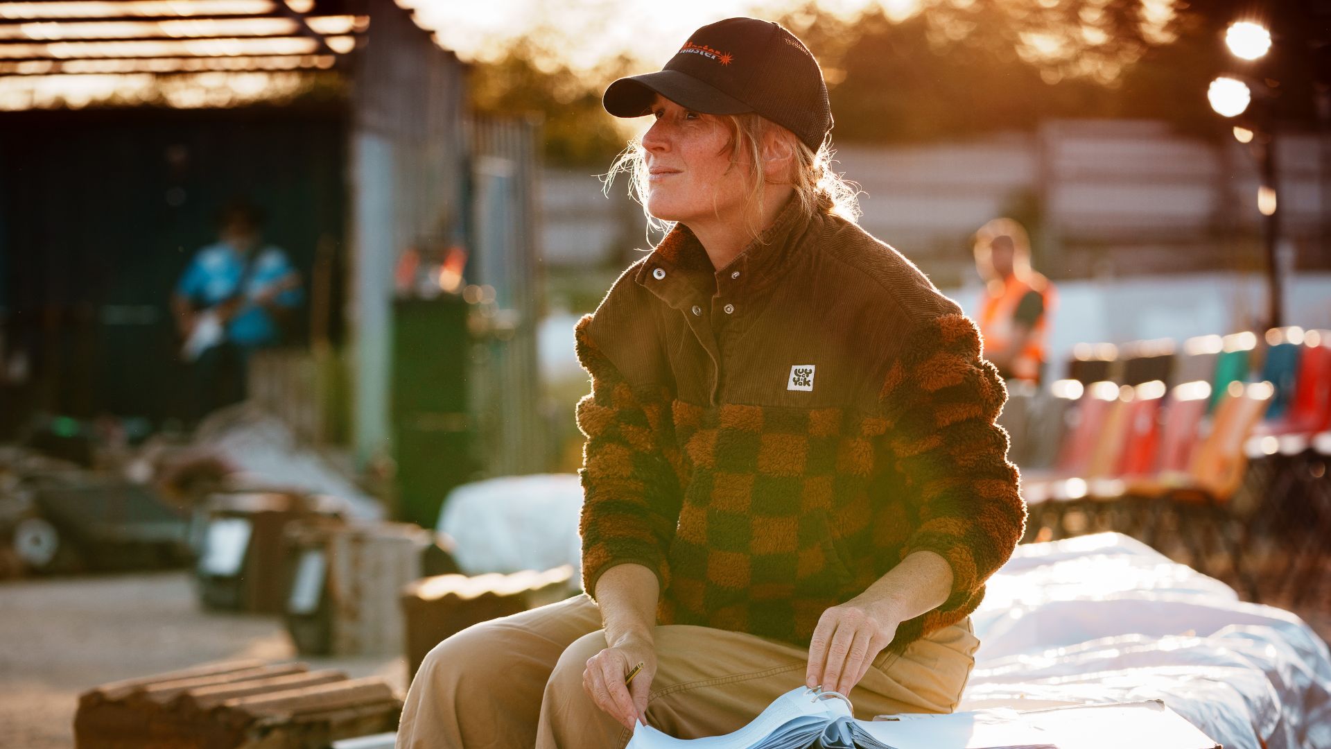 Kyla Goodey is sitting on hay bales directing Wildwork's show 'The Kneebone Cadillac'. It's golden hour and Kyla is wearing a baseball cap and checkered fleece.
